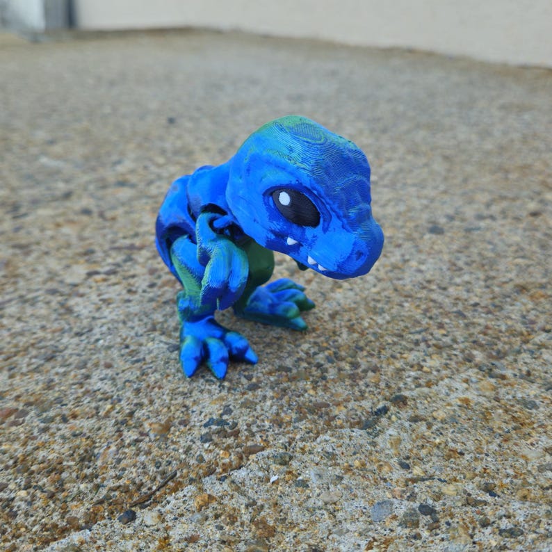May include: A small blue and green dinosaur figurine with a toothy grin. The dinosaur has large black eyes and is posed in a crouching position.