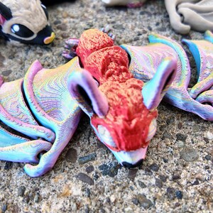 May include: A small, red, 3D printed bat with iridescent wings. The bat has a detailed, textured surface and is posed with its wings spread out.
