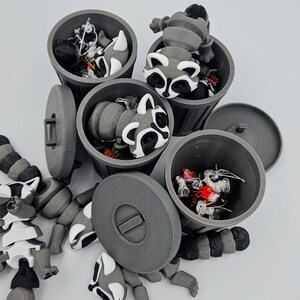 May include: A collection of gray 3D-printed trash cans with lids, each containing a raccoon figurine. The raccoons have black, white, and gray markings. One articulated raccoon figurine is partially disassembled nearby.