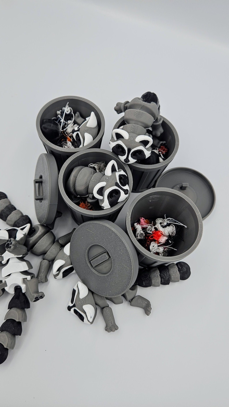 May include: A collection of gray 3D-printed trash cans with lids, each containing a raccoon figurine. The raccoons have black, white, and gray markings. One articulated raccoon figurine is partially disassembled nearby.