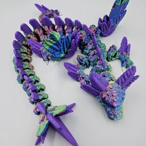 May include: A 3D printed dragon figurine with a purple and iridescent finish. The dragon is curled up in a coil with its head raised and its wings spread out. The dragon's scales are detailed and textured.