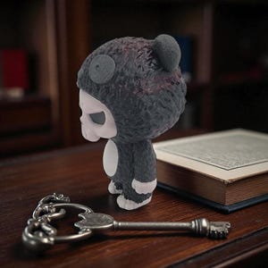 May include: A dark gray figurine in a bear suit with a skull-like face. The figurine has a small, round ear on top of its head and white accents on its chest and feet. A silver key lies on the wooden surface in front of the figurine.