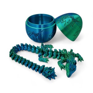 May include: A teal and green 3D printed dragon toy. The dragon is made up of multiple pieces that can be assembled. The dragon is in a closed egg-shaped container with a unicorn design.