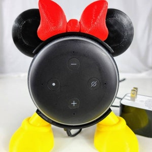 Puede incluir: Un altavoz inteligente Amazon Echo Dot negro con un soporte de Minnie Mouse impreso en 3D. El soporte tiene orejas rojas con un lazo rojo, una cara negra y pies amarillos.