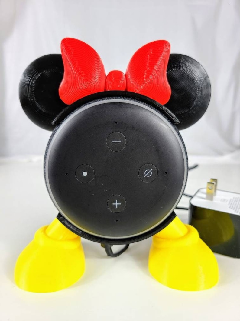 Puede incluir: Un altavoz inteligente Amazon Echo Dot negro con un soporte de Minnie Mouse impreso en 3D. El soporte tiene orejas rojas con un lazo rojo, una cara negra y pies amarillos.
