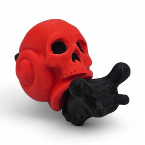 May include: A red skull sculpture with a black hand reaching out. The skull has hollow eye sockets and a detailed texture. The black hand is pointing forward, creating a striking contrast against the red skull.