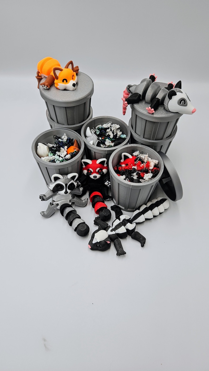 May include: A collection of miniature gray trash cans, each filled with colorful debris and topped with various animal figurines. The animals include a fox, opossum, raccoon, red panda, and skunk. The figurines are in various poses.