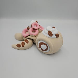 Pode incluir: Uma escultura extravagante de uma tartaruga com um design tem&aacute;tico de caf&eacute;. A carapa&ccedil;a da tartaruga &eacute; adornada com mini-rosquinhas rosa, enquanto sua cabe&ccedil;a apresenta um design de x&iacute;cara de caf&eacute; com chantilly e gr&atilde;os de caf&eacute;. A escultura &eacute; feita de argila marrom e bege.
