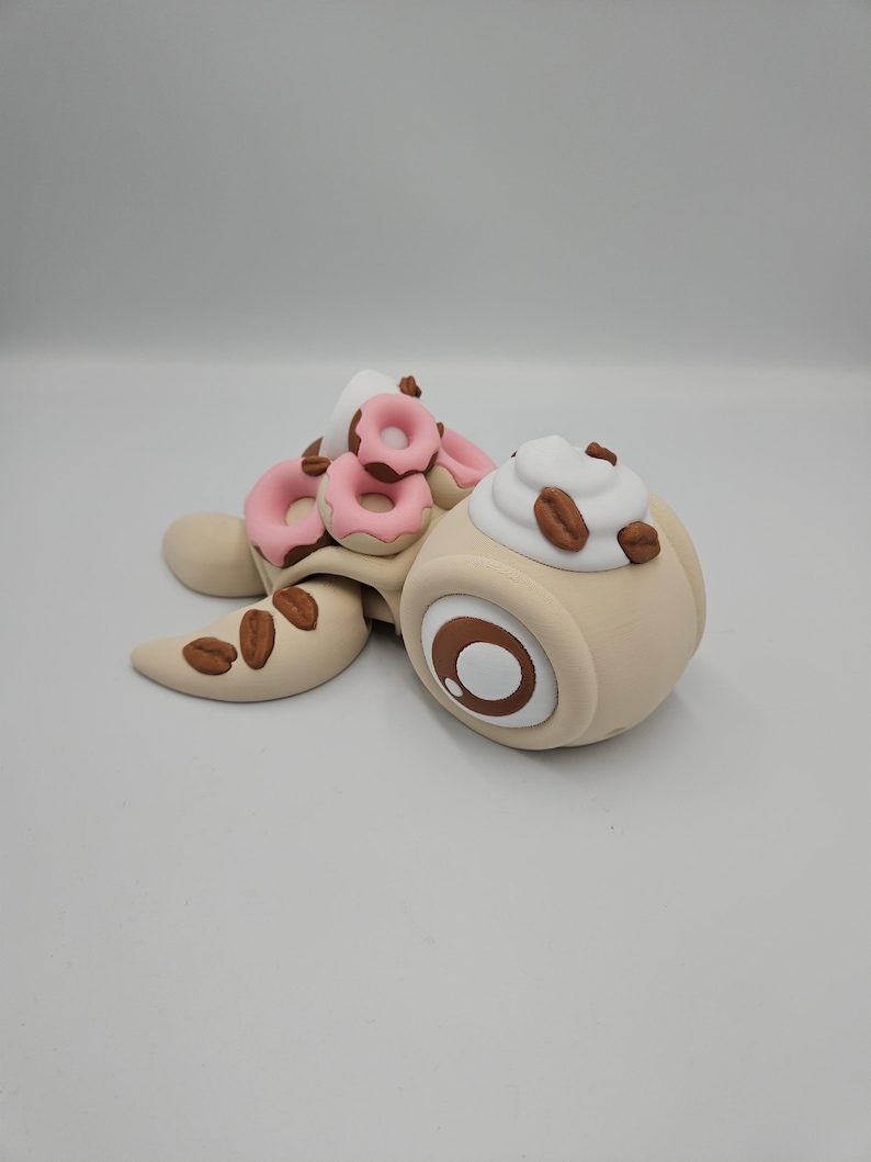 Pode incluir: Uma escultura extravagante de uma tartaruga com um design tem&aacute;tico de caf&eacute;. A carapa&ccedil;a da tartaruga &eacute; adornada com mini-rosquinhas rosa, enquanto sua cabe&ccedil;a apresenta um design de x&iacute;cara de caf&eacute; com chantilly e gr&atilde;os de caf&eacute;. A escultura &eacute; feita de argila marrom e bege.