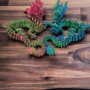 May include: Four articulated dragon figurines in vibrant colors: pink, green, and blue. The dragons are posed on a wooden surface, showcasing their detailed, scaled bodies and spiky features. These decorative figurines are a unique addition to any collection.