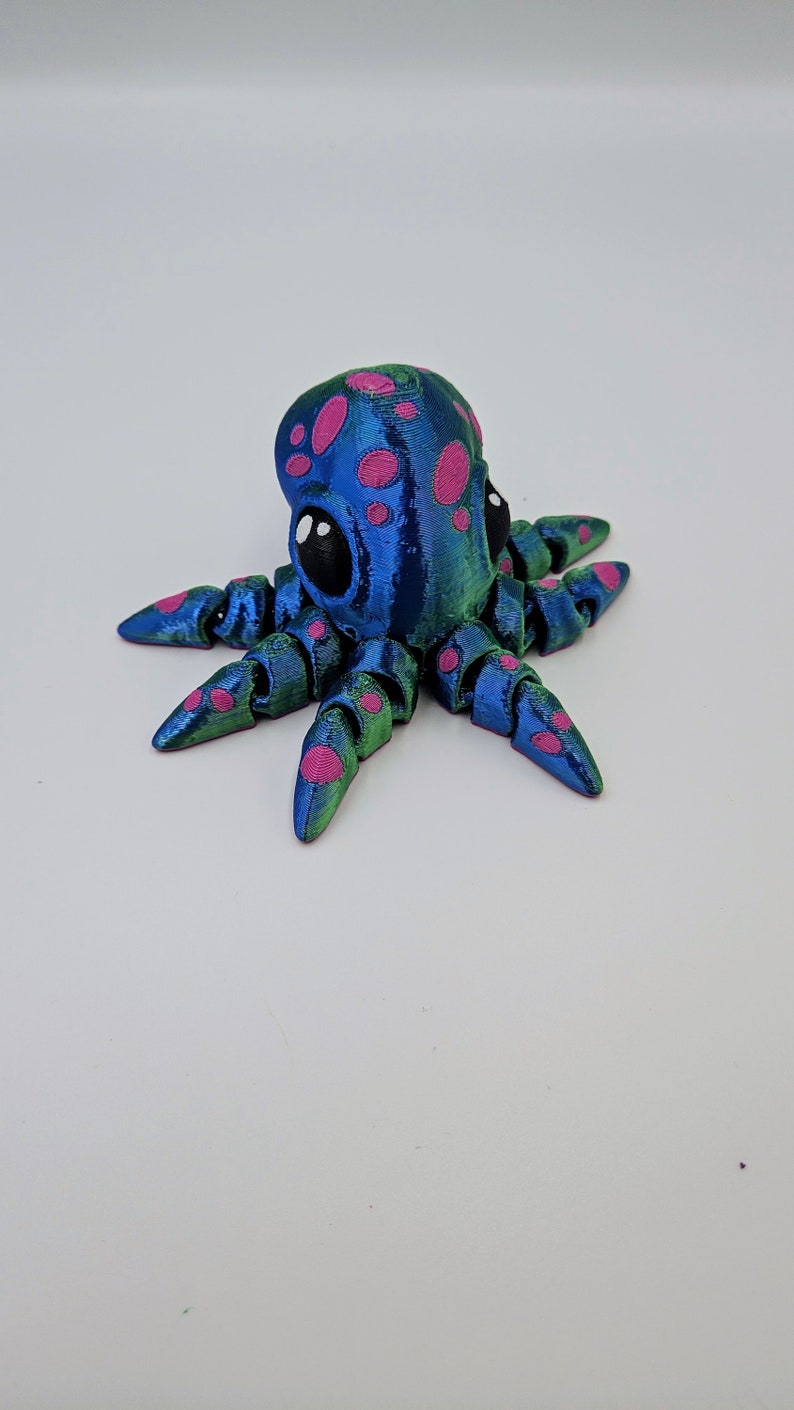 Puede incluir: Una peque&ntilde;a figura de pulpo azul y verde con lunares rosas y ojos negros. El pulpo tiene ocho tent&aacute;culos.