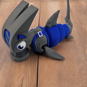 May include: A 3D printed gray and blue toy shark with a cartoonish expression. The shark has a blue body and gray fins, tail, and head. The shark's eyes are white with black pupils.