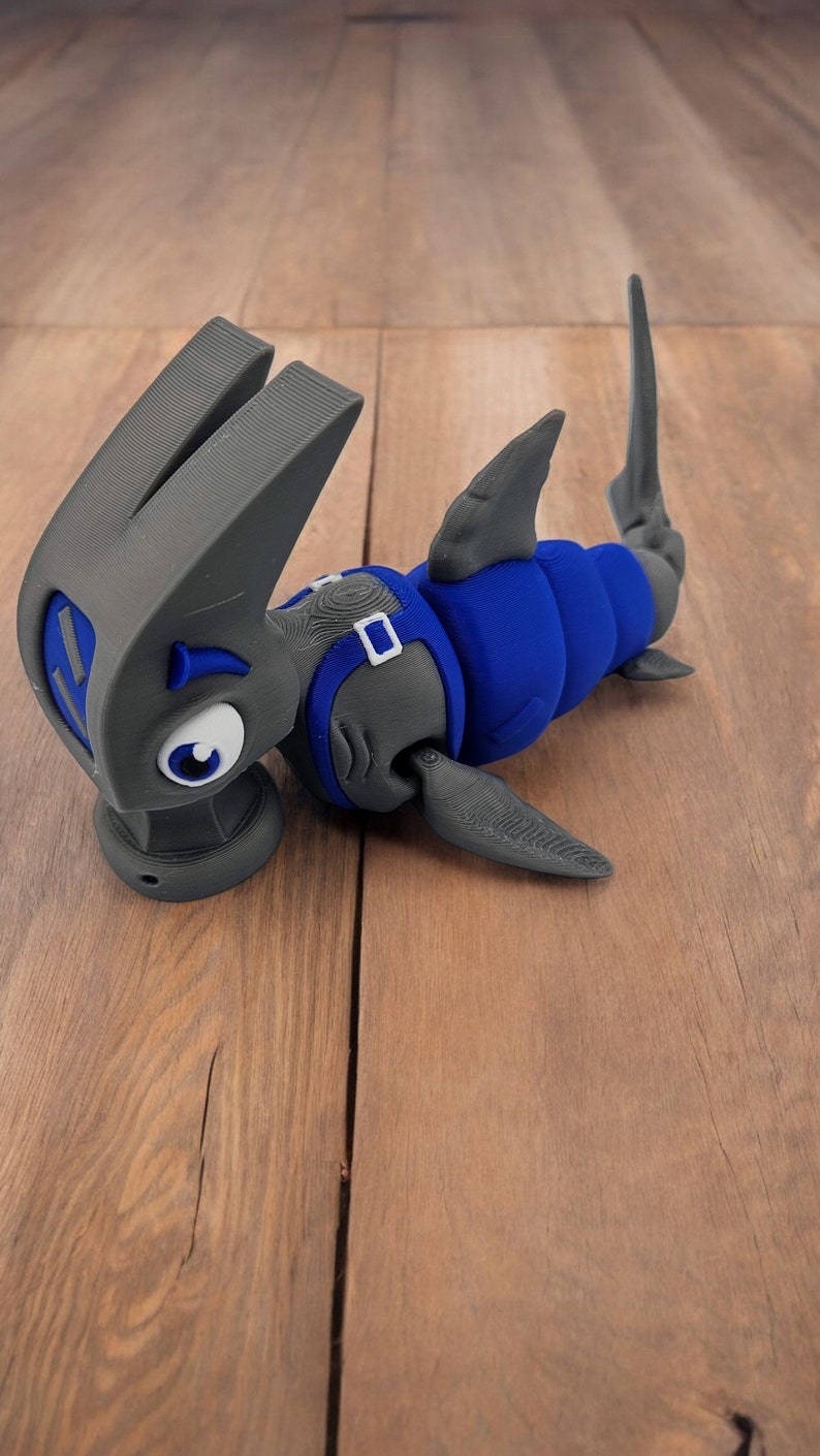 May include: A 3D printed gray and blue toy shark with a cartoonish expression. The shark has a blue body and gray fins, tail, and head. The shark's eyes are white with black pupils.