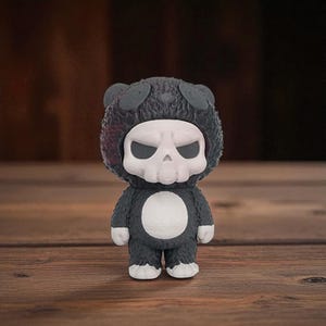 May include: A small, dark gray figurine of a character in a bear suit with a skull-shaped head. The skull is white with black eye sockets. The bear suit has a white belly and white feet. The figurine is standing on a wooden surface.