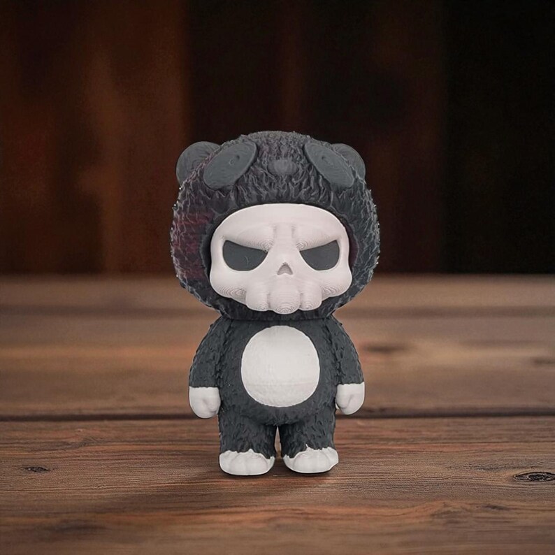 May include: A small, dark gray figurine of a character in a bear suit with a skull-shaped head. The skull is white with black eye sockets. The bear suit has a white belly and white feet. The figurine is standing on a wooden surface.