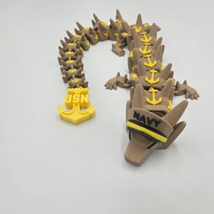 Pode incluir: Um brinquedo de drag&atilde;o articulado marrom e amarelo. A cabe&ccedil;a mostra a palavra "NAVY" e a cauda mostra "USN". O drag&atilde;o &eacute; feito de m&uacute;ltiplos segmentos e apresenta detalhes de &acirc;ncora.