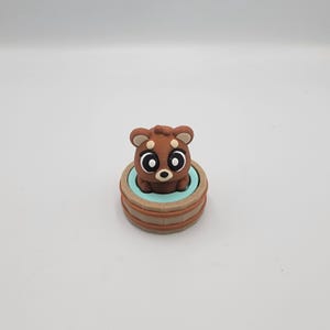 May include: A brown bear figurine with large, expressive eyes sits inside a small, light blue tub. The tub is surrounded by a wooden-textured border. The bear has a cute, cartoonish design.
