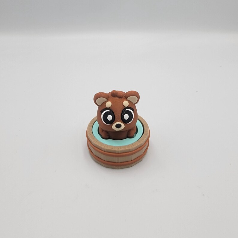 May include: A brown bear figurine with large, expressive eyes sits inside a small, light blue tub. The tub is surrounded by a wooden-textured border. The bear has a cute, cartoonish design.