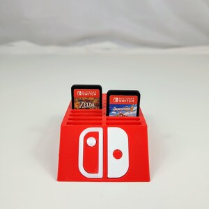 Może przedstawiać: Czerwony plastikowy stojak na karty do gier Nintendo Switch z białym logo Nintendo Switch. Stojak ma miejsca na 12 kart do gier. W stojaku znajdują się dwie karty do gier, jedna do gry "The Legend of Zelda: Breath of the Wild" i jedna do gry "Overcooked! 2".