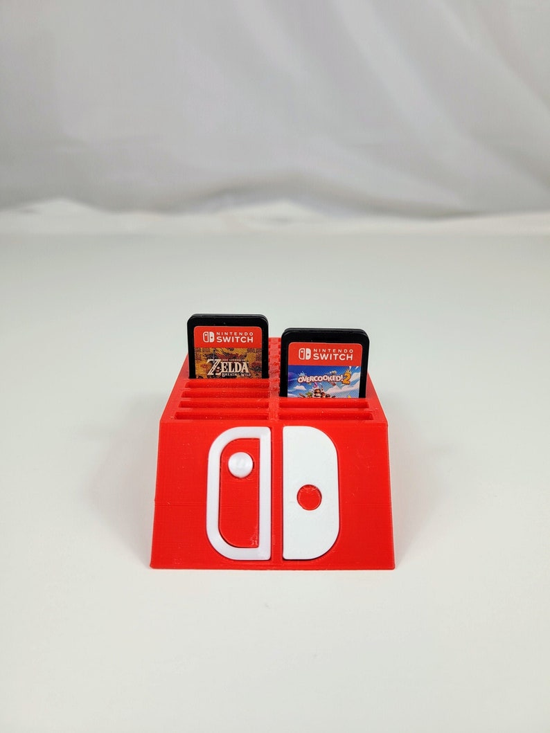 Może przedstawiać: Czerwony plastikowy stojak na karty do gier Nintendo Switch z białym logo Nintendo Switch. Stojak ma miejsca na 12 kart do gier. W stojaku znajdują się dwie karty do gier, jedna do gry "The Legend of Zelda: Breath of the Wild" i jedna do gry "Overcooked! 2".