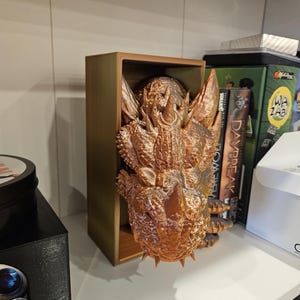 May include: A bronze-colored dragon head sculpture is displayed in a wooden box. The dragon has a textured surface and sharp teeth. The sculpture is a decorative item for a home or office.