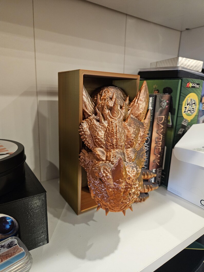 May include: A bronze-colored dragon head sculpture is displayed in a wooden box. The dragon has a textured surface and sharp teeth. The sculpture is a decorative item for a home or office.