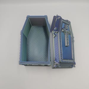 May include: An open, decorative coffin with a cross on the lid. The coffin is blue and green with ornate detailing. The interior is a lighter shade of blue. The coffin is likely a decorative item or a unique storage box.