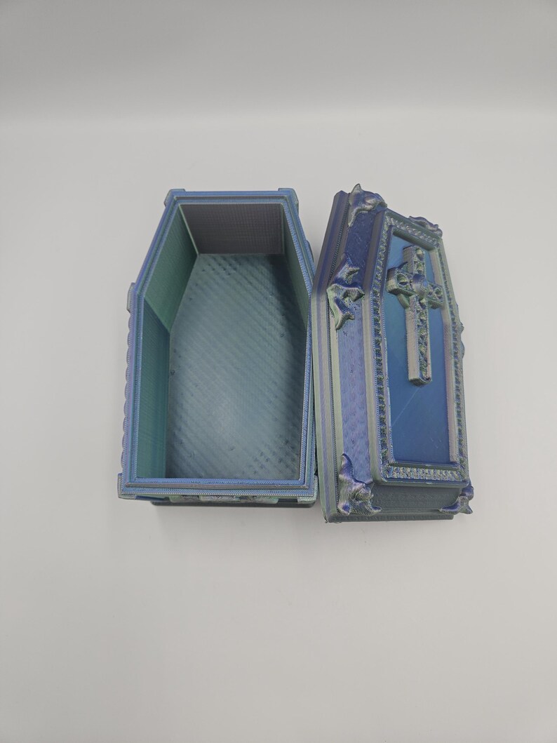 May include: An open, decorative coffin with a cross on the lid. The coffin is blue and green with ornate detailing. The interior is a lighter shade of blue. The coffin is likely a decorative item or a unique storage box.