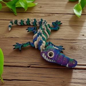 Puede incluir: Una colorida escultura de caimán articulada hecha de arcilla. El caimán presenta una cabeza morada, azul y verde con dientes blancos y un ojo grande. El cuerpo es una mezcla de verde, azul y blanco, con una columna vertebral segmentada.