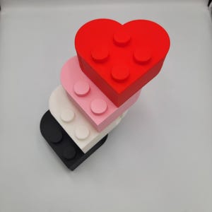 May include: A stack of heart-shaped building blocks in red, pink, white, and black. Each block has circular studs on the top, resembling toy bricks. The blocks are arranged in a slightly offset, layered design.
