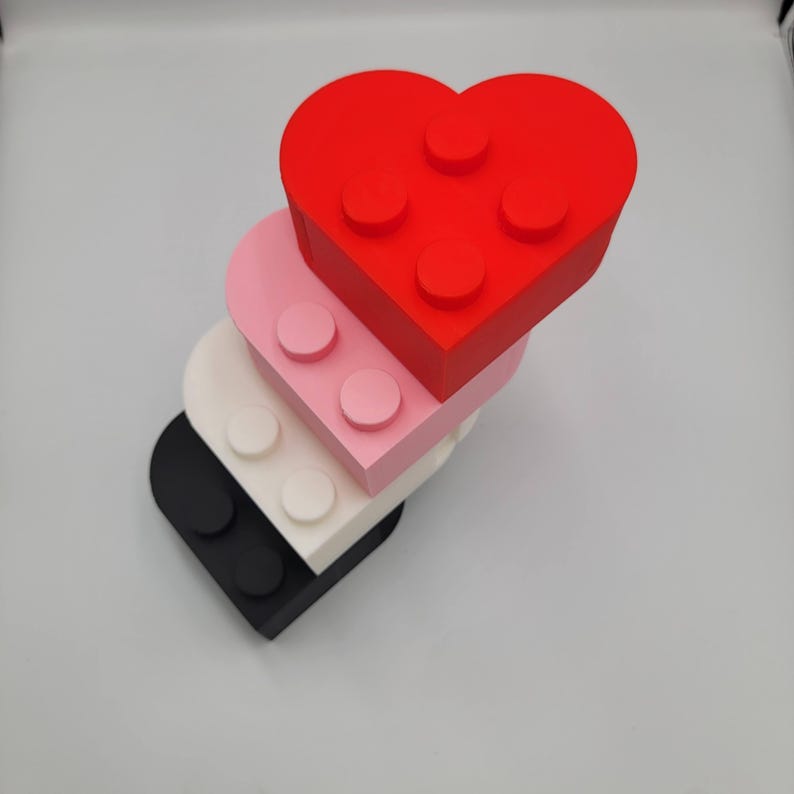 May include: A stack of heart-shaped building blocks in red, pink, white, and black. Each block has circular studs on the top, resembling toy bricks. The blocks are arranged in a slightly offset, layered design.