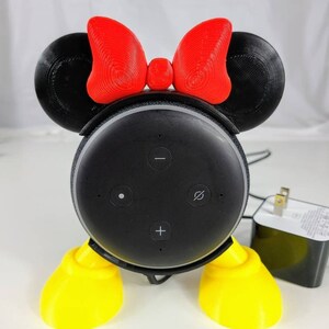Puede incluir: Un altavoz inteligente Amazon Echo Dot negro con un soporte Minnie Mouse impreso en 3D. El soporte tiene orejas rojas con un lazo rojo, zapatos amarillos y pies negros.