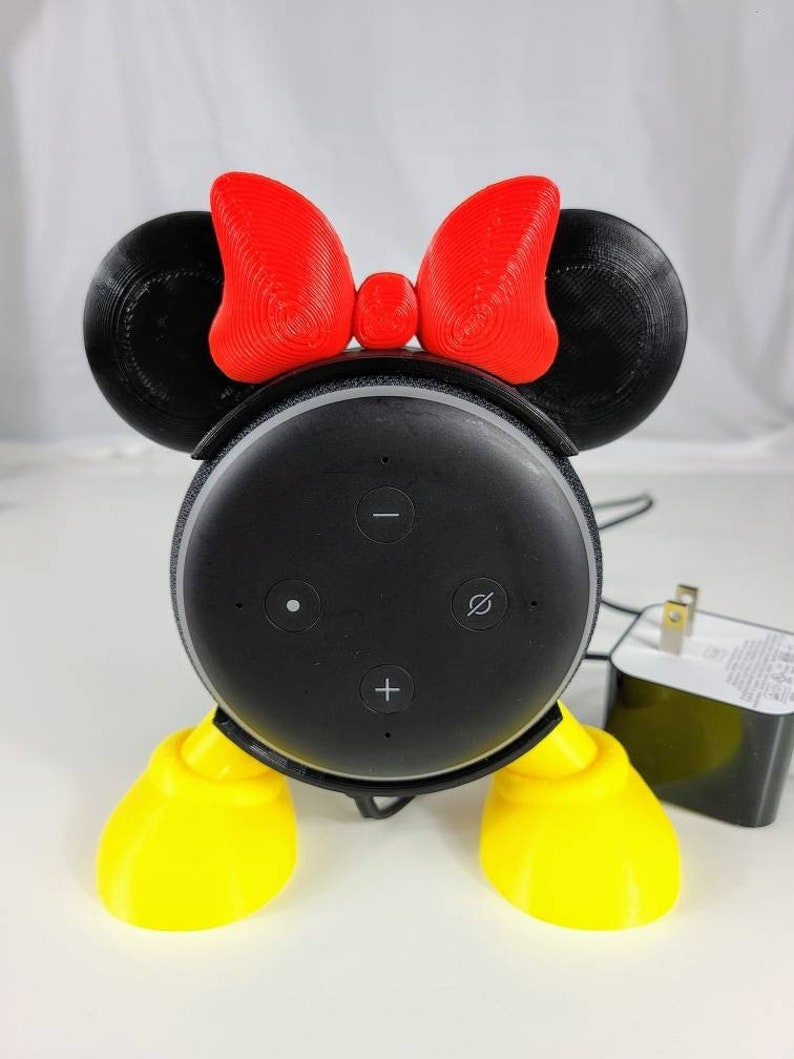 Puede incluir: Un altavoz inteligente Amazon Echo Dot negro con un soporte Minnie Mouse impreso en 3D. El soporte tiene orejas rojas con un lazo rojo, zapatos amarillos y pies negros.