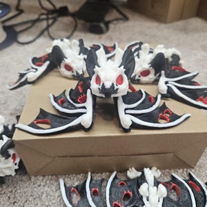 May include: A collection of small, decorative bat figurines. Each bat features a white head and body, black wings with red accents, and red eyes. The bats are arranged on a cardboard surface.