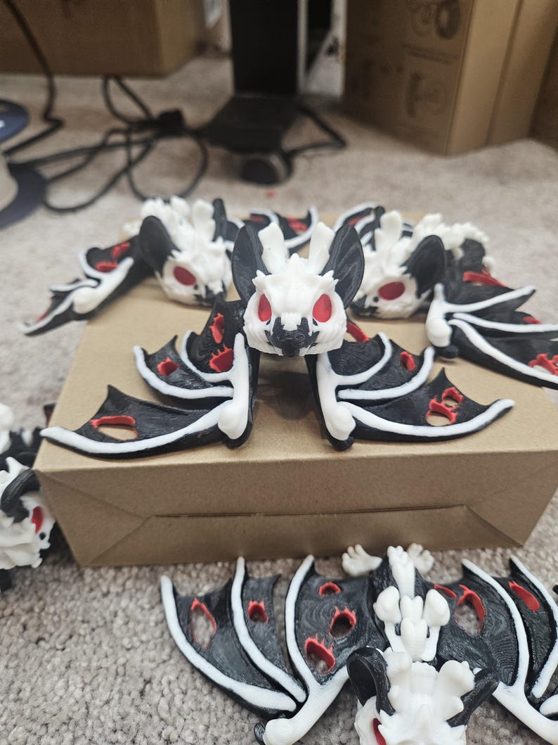 May include: A collection of small, decorative bat figurines. Each bat features a white head and body, black wings with red accents, and red eyes. The bats are arranged on a cardboard surface.