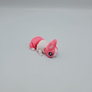 K&ouml;nnte beinhalten: Ein rosa-wei&szlig;es, artikuliertes Spielzeugkaninchen mit schwarzen Augen. Das Kaninchen liegt auf dem Bauch.