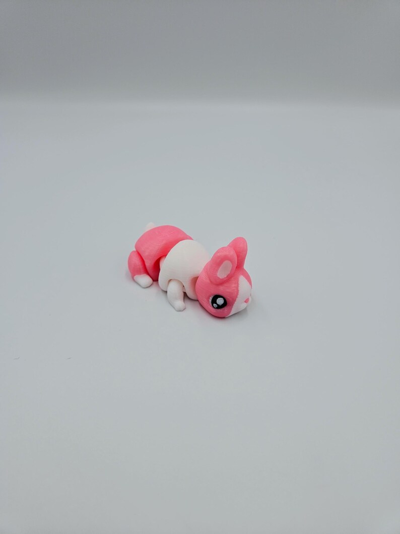 K&ouml;nnte beinhalten: Ein rosa-wei&szlig;es, artikuliertes Spielzeugkaninchen mit schwarzen Augen. Das Kaninchen liegt auf dem Bauch.