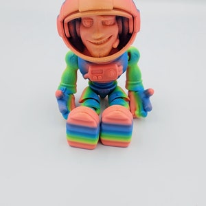 May include: A small, colorful astronaut figurine with a rainbow gradient paint job. The astronaut is sitting with its legs crossed and has a friendly expression on its face.