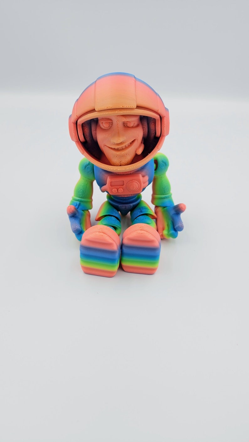 May include: A small, colorful astronaut figurine with a rainbow gradient paint job. The astronaut is sitting with its legs crossed and has a friendly expression on its face.
