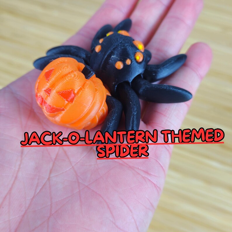 Puede incluir: Un juguete de ara&ntilde;a negro y naranja con una cabeza de calabaza. La ara&ntilde;a tiene ocho patas y un cuerpo negro con ojos naranjas. La cabeza de calabaza tiene una cara tallada con una amplia sonrisa y dos tri&aacute;ngulos para los ojos.