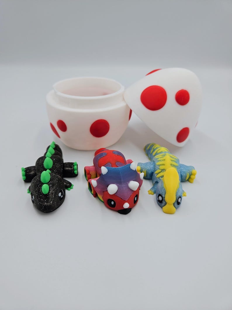 May include: A set of three small dinosaur toys, including a black and green dinosaur, a multi-colored dinosaur, and a blue and yellow dinosaur. The dinosaurs are sitting inside a white egg-shaped container with red polka dots.