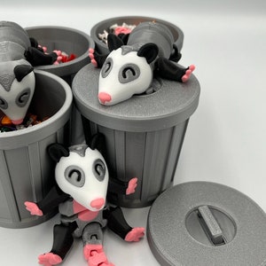 Op de afbeelding: Een verzameling grijze 3D-geprinte vuilnisbakken met opossum beeldjes. De opossums hebben witte gezichten, roze neuzen en voeten, en gelede roze staarten. Sommige opossums zitten in de vuilnisbakken.