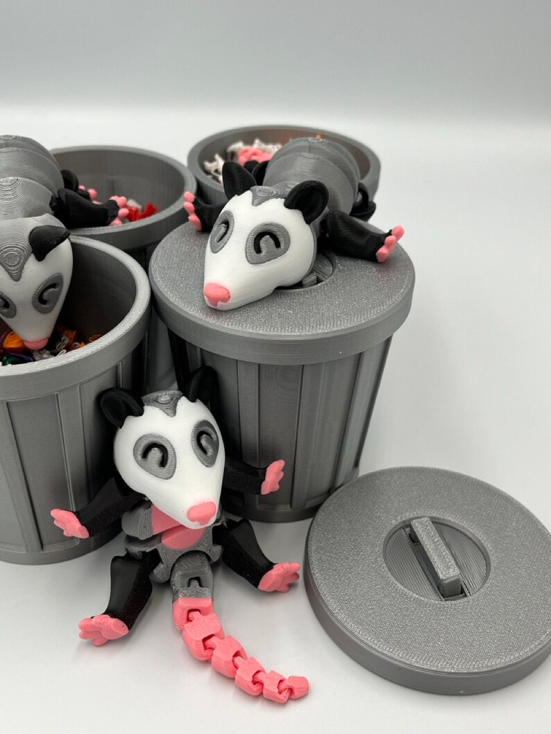 Op de afbeelding: Een verzameling grijze 3D-geprinte vuilnisbakken met opossum beeldjes. De opossums hebben witte gezichten, roze neuzen en voeten, en gelede roze staarten. Sommige opossums zitten in de vuilnisbakken.
