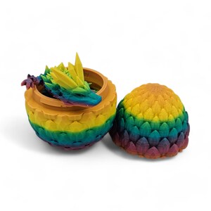 May include: A 3D-printed dragon egg, open to reveal a colorful dragon figurine. The egg is yellow, green, and blue, with a matching lid. The dragon is multi-colored, with yellow, green, blue, and purple hues.