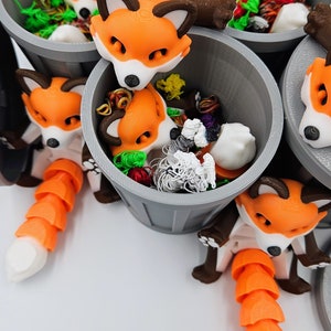 May include: A collection of orange and white fox figurines with brown accents, each nestled in a gray bucket filled with colorful craft supplies. The foxes have segmented tails and are arranged against a white background.