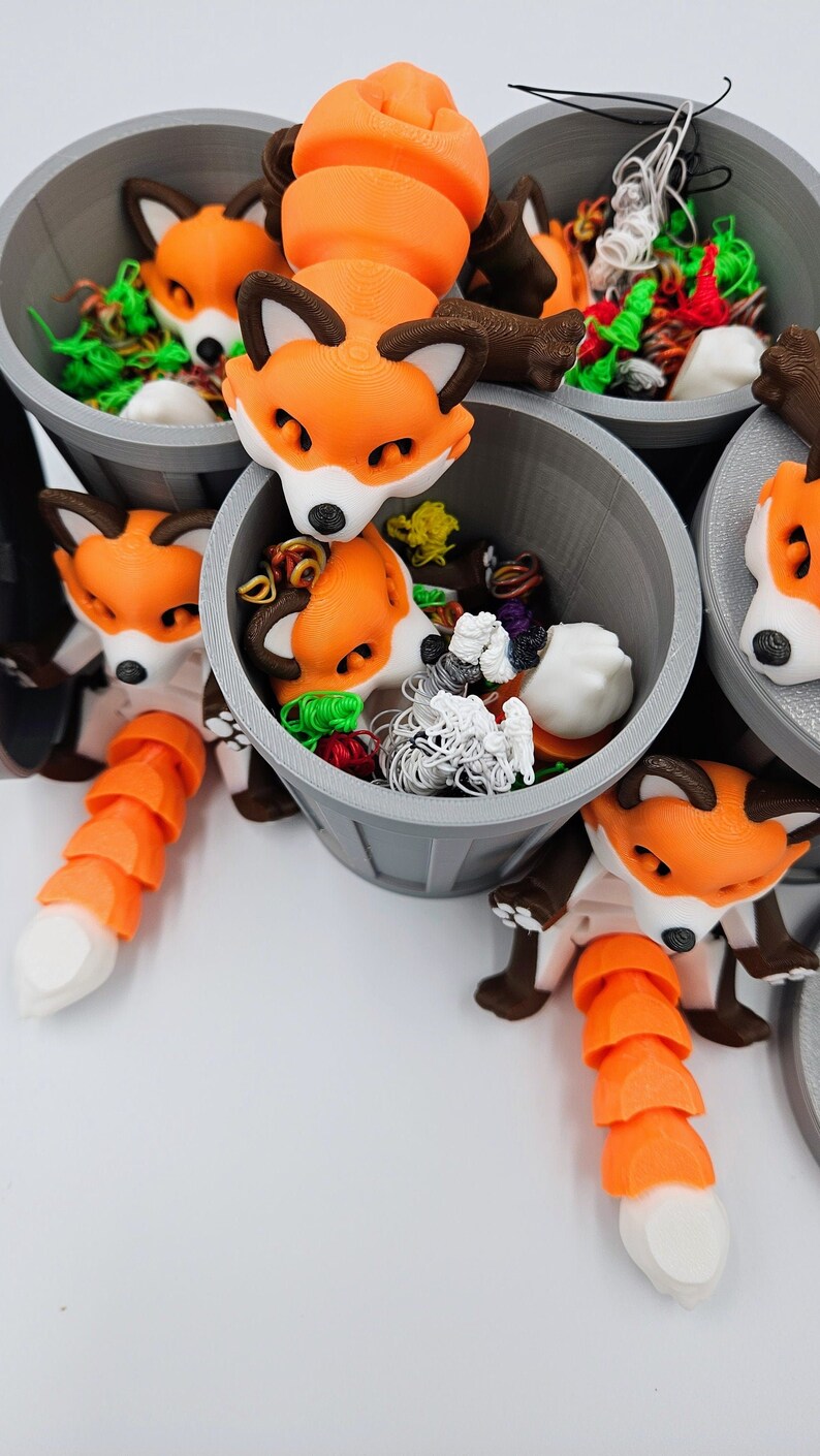 May include: A collection of orange and white fox figurines with brown accents, each nestled in a gray bucket filled with colorful craft supplies. The foxes have segmented tails and are arranged against a white background.