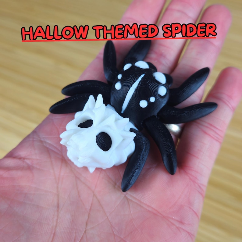 Puede incluir: Una ara&ntilde;a impresa en 3D en blanco y negro con un cuerpo en forma de calavera. La ara&ntilde;a tiene un cuerpo negro con puntos blancos y una calavera blanca con ojos negros. HALLOW THEMED SPIDER est&aacute; escrito en letras rojas en la parte superior de la imagen.