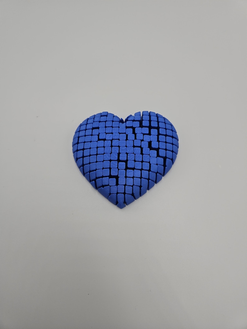 May include: A blue heart-shaped object composed of numerous small, square blocks. The heart has a textured appearance and is set against a plain, light background. The design gives a 3D effect.