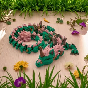 May include: A handcrafted, green and pink dragon figurine with intricate details. The dragon is coiled, with a detailed head and spiky accents. The figurine is surrounded by greenery and flowers, creating a whimsical, fantasy-themed display.