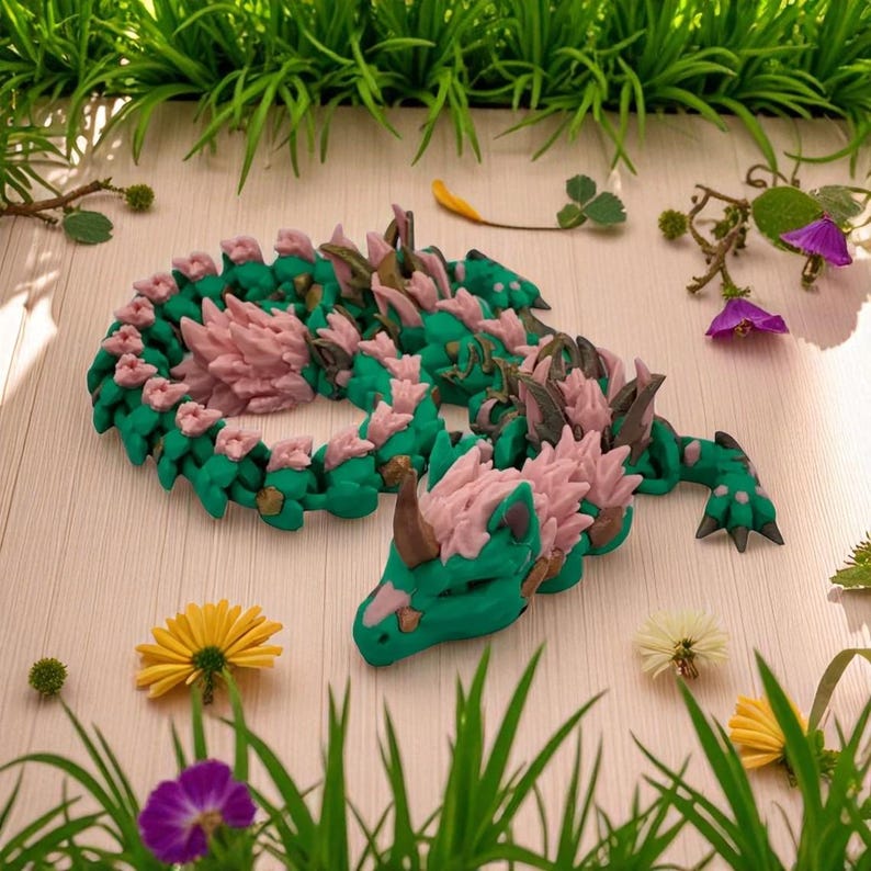 May include: A handcrafted, green and pink dragon figurine with intricate details. The dragon is coiled, with a detailed head and spiky accents. The figurine is surrounded by greenery and flowers, creating a whimsical, fantasy-themed display.